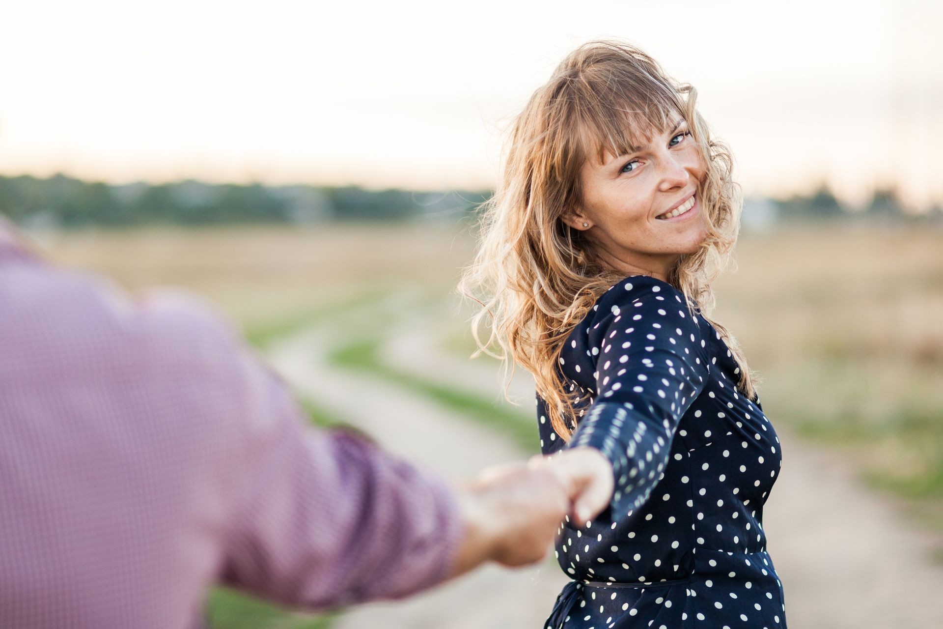 Traveling together. Valentine's day. Follow me. Young blond woman holding boyfriend's hand walking in the field on sunset